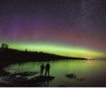 Two people stand hand-in-hand at the edge of a peaceful lake, illuminated by the soft green and violet hues of the Northern Lights.