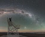 A lone person sits atop a metal observation staircase under a vivid, star-studded Milky Way sky in a barren, desert-like landscape.