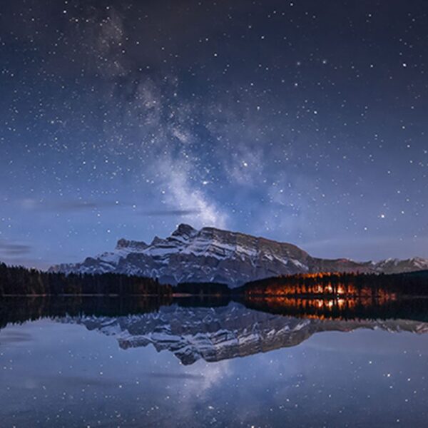 A panoramic nightscape featuring the Milky Way stretching across a still lake, with stars shimmering above a pine forest and snow-covered mountain.