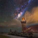 A lighthouse glowing under a breathtaking night sky filled with stars and the Milky Way, surrounded by glowing clouds