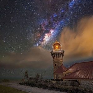 A lighthouse glowing under a breathtaking night sky filled with stars and the Milky Way, surrounded by glowing clouds