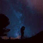 Silhouetted couple sharing a kiss under a stunning Milky Way sky with a lone tree framing the cosmic backdrop.
