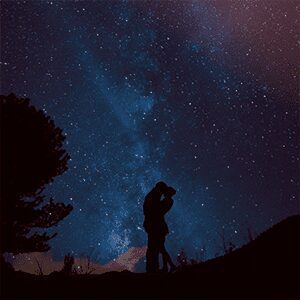 Silhouetted couple sharing a kiss under a stunning Milky Way sky with a lone tree framing the cosmic backdrop.