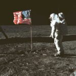 An astronaut stands beside the American flag on the surface of the Moon, with visible boot prints in the lunar dust beneath a black space backdrop.