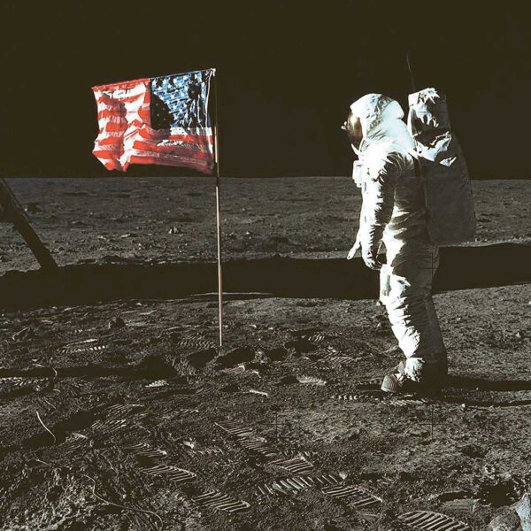 An astronaut stands beside the American flag on the surface of the Moon, with visible boot prints in the lunar dust beneath a black space backdrop.