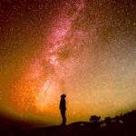 Silhouette of a person standing under a vivid red-orange aurora borealis sky filled with stars