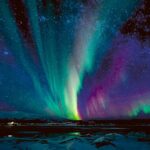 A vibrant aurora borealis in shades of green, turquoise, and purple dancing above a snow-covered landscape and icy lake under a star-filled sky