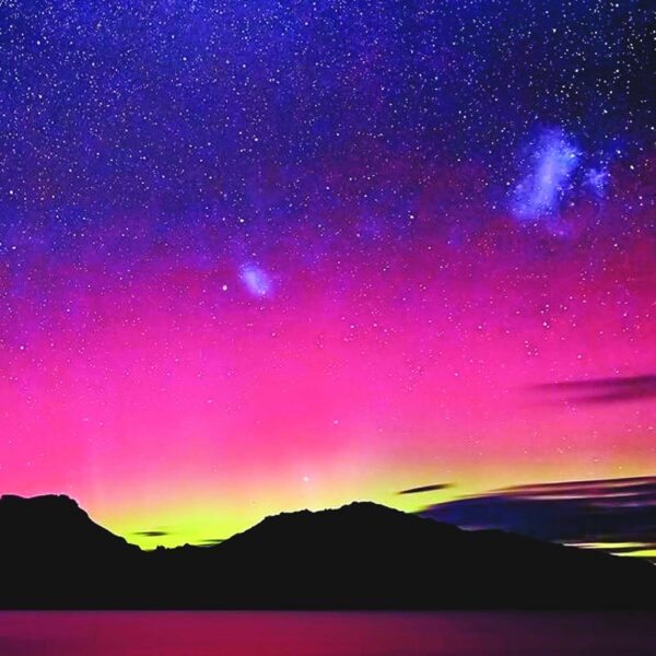 A surreal night sky filled with pink, purple, and electric blue hues from the aurora borealis above a dark mountain range and still lake