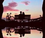 A couple sitting on a bench at sunset with a bicycle nearby, reflected perfectly in still water