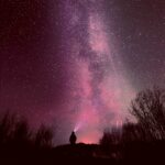 A person stands silhouetted against a starry sky with the northern lights in vibrant pinks and purples, casting a magical glow over the landscape."