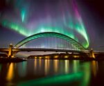 A vibrant display of the northern lights illuminates the sky above the iconic Newcastle bridge, reflecting in the calm waters below, with colors of green, pink, and purple swirling across the night sky.