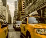 A classic yellow NYC taxi driving through a busy street lined with skyscrapers and urban traffic