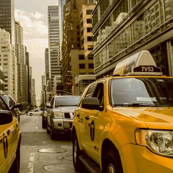 A classic yellow NYC taxi driving through a busy street lined with skyscrapers and urban traffic