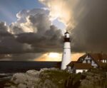 A lighthouse stands strong on rocky cliffs as storm clouds part and sunlight streams through over the ocean