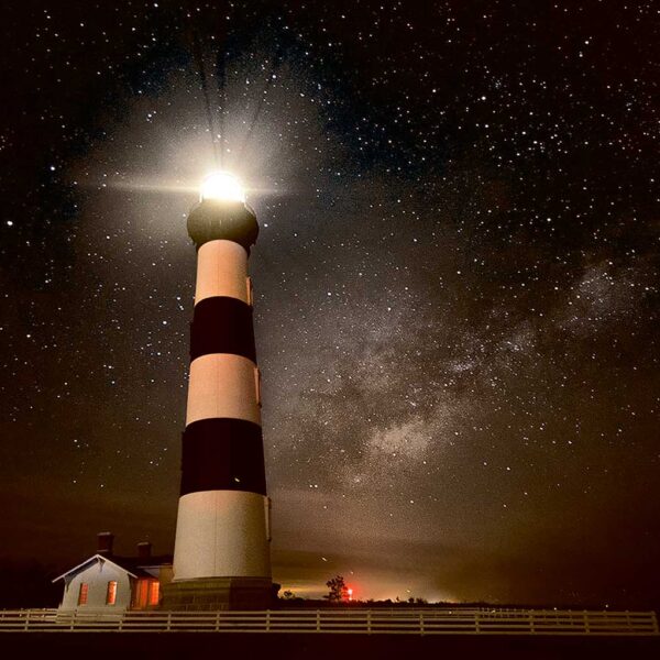 A tall black-and-white striped lighthouse glowing at night under a star-filled sky and the Milky Way