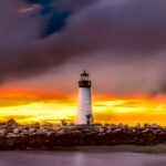 A lighthouse standing on rocky shore illuminated by a dramatic sunset with purple and golden clouds